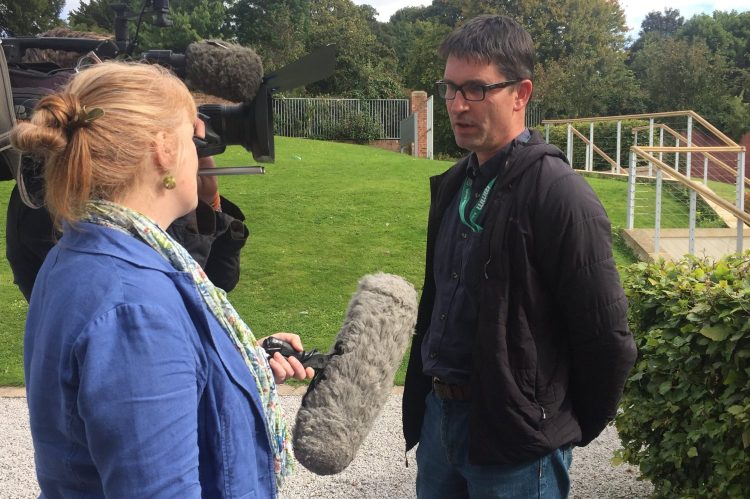 A woman interviewing a man, using a large microphone. They are being filed by a man (hidden) with a TV camera