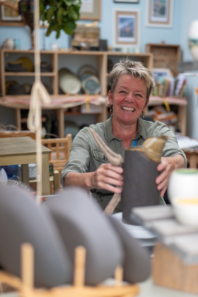 Georgi Gilpin in her studio. her hands are on a jug in front of her. In the background you can see the contents of her studio, out of focus.